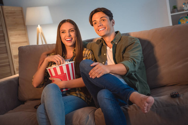 Photo of cheerful cute young couple wear casual shirts smiling sitting couch watching tv laughing eating pop corn indoors house room - Φωτογραφία, εικόνα