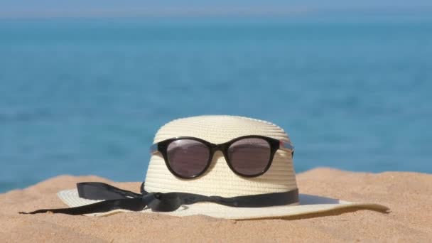 Closeup of yellow straw hat and black protective sunglasses on sandy beach at tropical seaside on warm sunny day. Summer vacation concept - Footage, Video
