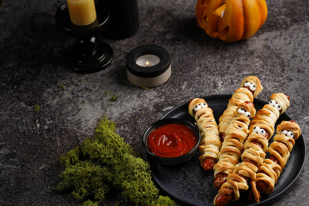 Snack de fiesta de Halloween lindo: salchicha salchicha con ojos de azúcar envueltos en rayas de masa que se asemejan a momias en un plato negro con salsa de tomate - Foto, Imagen
