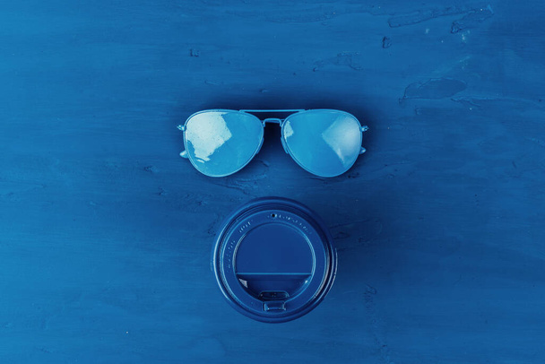 Top view of takeaway coffee cup with aviator sunglasses on classic blue background - Foto, afbeelding