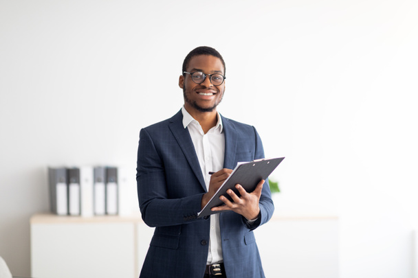 Boldog fiatal fekete férfi pszichológus írás vágólapon, nézi a kamera és mosolyog a modern irodában - Fotó, kép
