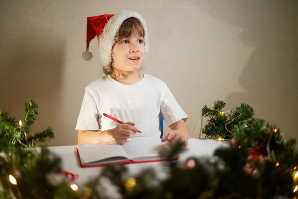 girl child in a white T-shirt with a red pen and a New Year's cap on his head writes dear Santa a letter at the table in anticipation of the birth. wish list of gifts for the new year - Foto, afbeelding