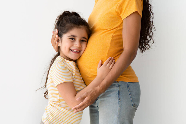 Портрет очаровательной маленькой девочки, обнимающей беременных матерей животом, крупным планом - Фото, изображение