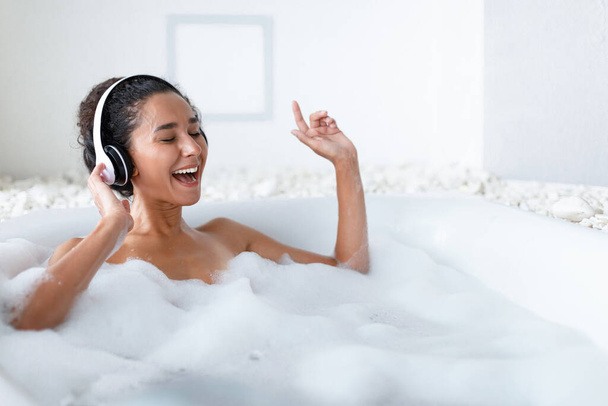Jovem atraente usando fones de ouvido e cantando enquanto estava deitado em banho quente em casa, espaço de cópia - Foto, Imagem