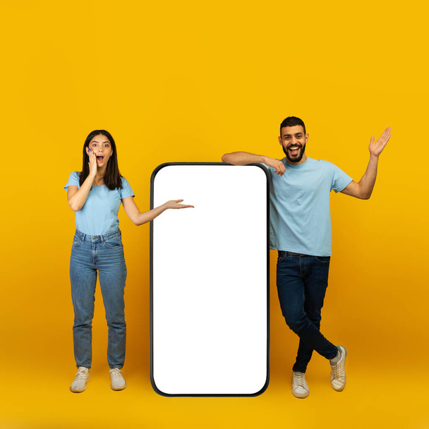 Şaşırmış Arap çift, sarı stüdyo arka planında boş ekranları olan akıllı telefonun yanında duruyor. - Fotoğraf, Görsel