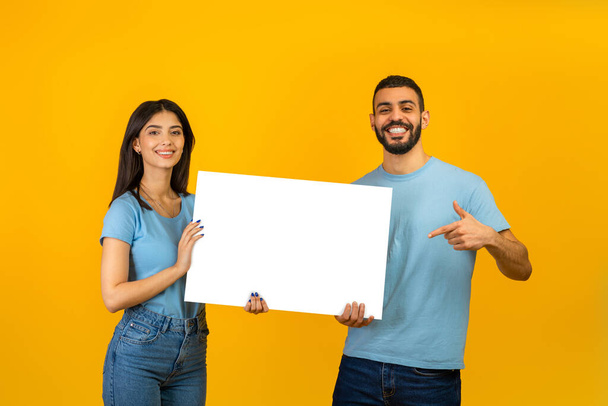 Portrait of happy arab couple holding white placard, man pointing at free space for advertising, yellow background - Фото, изображение
