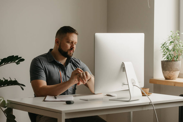 Un hombre trabaja remotamente en una computadora de escritorio. Un tipo con barba está moviendo su reloj inteligente durante una videoconferencia en casa. Un profesor se prepara para una conferencia en línea. - Foto, Imagen