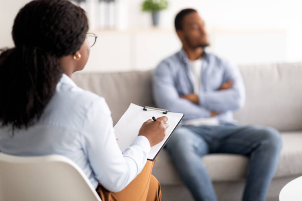 Молодой чернокожий мужчина с психическими проблемами проводит сеанс с психотерапевтом в медицинском кабинете, избирательный фокус - Фото, изображение
