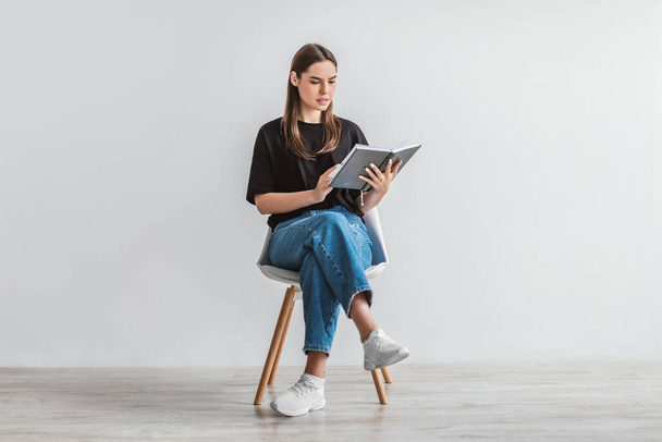 Serious young woman reading book, sitting on chair, getting ready for college exams against white wall, full length - Foto, Imagem