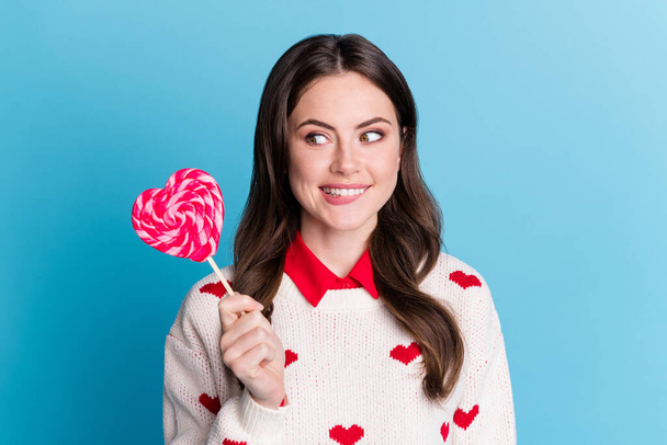 Photo of adorable tricky young lady wear hearts print sweater holding looking ready eat candy isolated blue color background - 写真・画像