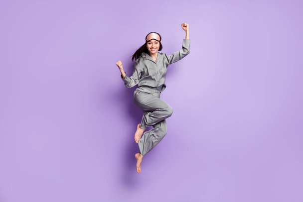 Teljes hossz testméret fotó lány jumping visel pizsama alvó maszk gesztikulál, mint a győztes elszigetelt pasztell ibolya szín háttér - Fotó, kép