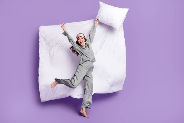 Full length body size photo of woman jumping keeping pillow in grey sleepwear gesturing like winner isolated pastel purple color background - Zdjęcie, obraz