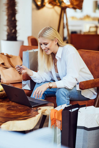 Μια νεαρή ξανθιά μοντέρνα γυναίκα κάθεται σε μια καφετέρια και πληκτρολογεί έναν αριθμό πιστωτικής κάρτας στο λάπτοπ της. Έχει σακούλες δίπλα στο πόδι της. Μια γυναίκα αγοράζει online. - Φωτογραφία, εικόνα