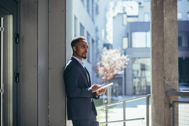 Young african american businessman in formal business suit standing working with tablet in hands on background modern office building outside. Man using smartphone or mobile phone outdoors city street - Foto, imagen