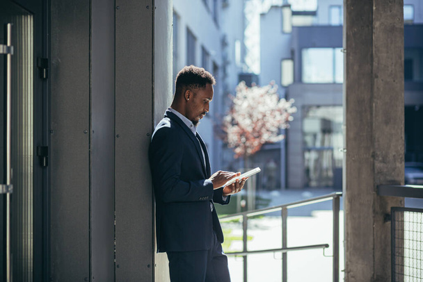 Young african american businessman in formal business suit standing working with tablet in hands on background modern office building outside. Man using smartphone or mobile phone outdoors city street - Foto, imagen