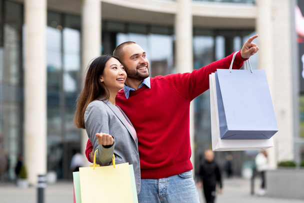 Pozitivní různorodý pár kráčející po ulici s papírovými taškami, ukazující na nový obchod nebo reklamu, nakupování společně - Fotografie, Obrázek