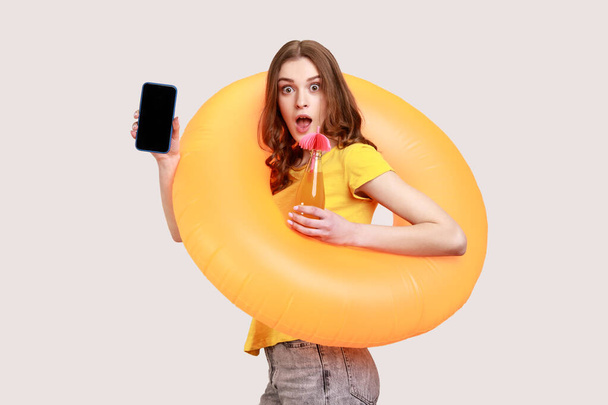 Amazed woman of young age in yellow T-shirt holding rubber ring, juice and looking at camera with shocked expression, showing blank smart phone screen. Indoor studio shot isolated on gray background. - Foto, Imagen