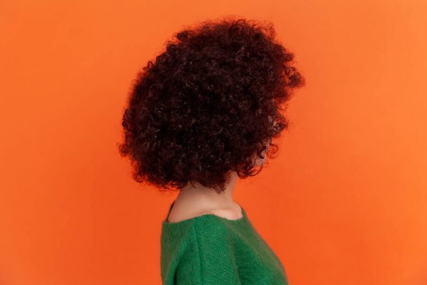 Side view of woman with Afro hairstyle wearing green casual style sweater showing her beautiful hair, advertising hairdresser or beauty salon. Indoor studio shot isolated on orange background. - Фото, изображение
