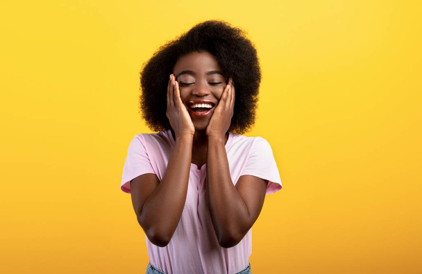 Szczęście i radość. Ładna Afroamerykanka trzymająca się za ręce na policzkach i uśmiechająca z zamkniętymi oczami, żółte tło - Zdjęcie, obraz