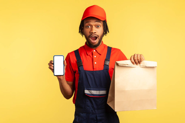 Wow, delivery service app! Deliveryman showing paper parcel and cellphone with mockup display for advertisement, looking shocked at camera. Indoor studio shot isolated on yellow background. - Foto, Bild