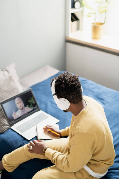 オンラインレッスン中にベッドに座っているヘッドフォンやホームウェアの若い学生 - 写真・画像