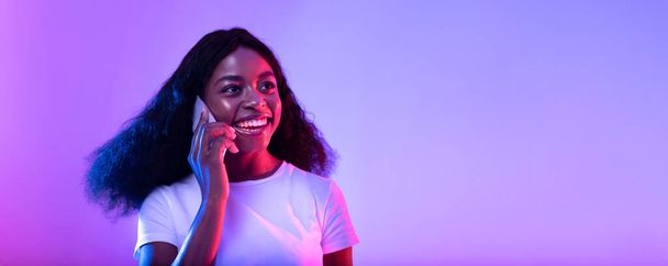 Mujer negra joven feliz hablando en el teléfono inteligente en luz de neón, diseño de pancartas con espacio vacío - Foto, imagen
