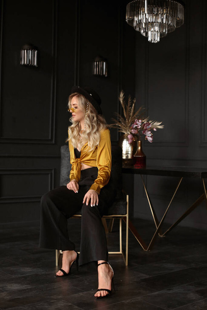 Confident model girl with long blond hair wearing fashionable outfit poses in dark luxurious interior - Foto, imagen