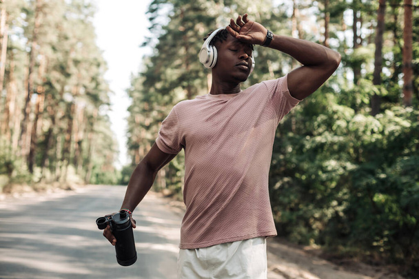 Young exhausted african man , runner resting after workout on a sunny morning, sportive man in headphones, outdoors on the road taking a break from jogging workout, athlete relaxing after workout - Photo, image