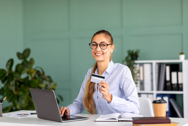 Retrato de mulher de negócios feliz fazendo compras on-line no computador portátil e segurando cartão de crédito, interior do escritório - Foto, Imagem