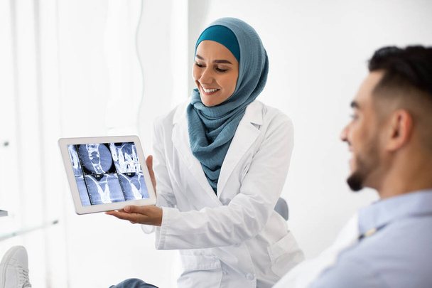 Muzułmanka Dentystka posiadająca cyfrowe tabletki, wyjaśniające proces leczenia męskiemu pacjentowi - Zdjęcie, obraz