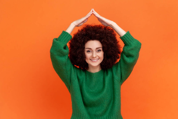 Woman with Afro hairstyle wearing green casual style sweater making house roof gesture and smiling, concept of life insurance, protection. Indoor studio shot isolated on orange background. - Photo, image
