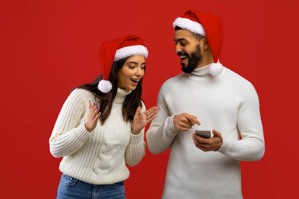 素晴らしいクリスマスのニュース。サンタの帽子で喜びに満ちたアラブの配偶者,メッセージを受信,妻にスマートフォンを示す男 - 写真・画像