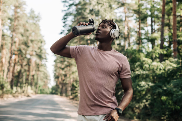 Sporty tired african american man , runner drinking water resting after workout, man relaxing after workout on city road in forest in nature - Fotó, kép