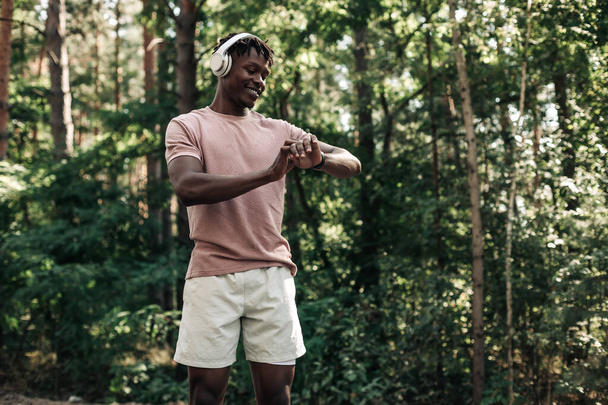 Corredor masculino, hombre americano que comprueba progreso de la aptitud en su smartwatch, hombre negro que usa la aplicación de la aptitud para seguir los resultados del entrenamiento mientras que camina en el bosque en la naturaleza - Foto, imagen