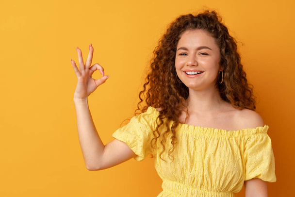 Young curly smiling woman showing ok sign against yellow background - 写真・画像