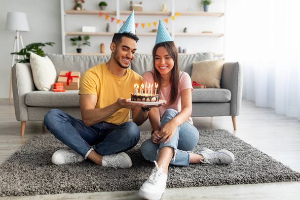Doğum günü pastasıyla yerde oturmuş, oturma odasında kutlama yapan neşeli şapkalı genç bir çift. - Fotoğraf, Görsel