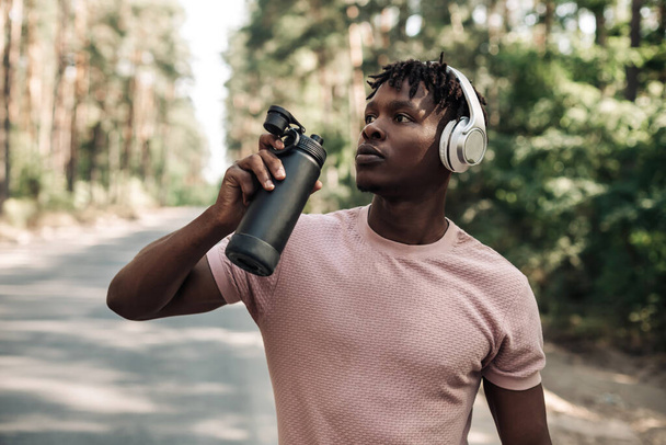 Sporty tired african american man , runner drinking water resting after workout, man relaxing after workout on city road in forest in nature - Foto, Imagen