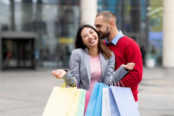 Liebender junger Kerl küsst seine lachende asiatische Freundin mit Einkaufstüten, drückt Zuneigung in der Nähe eines großen Einkaufszentrums aus, kopiert Raum - Foto, Bild