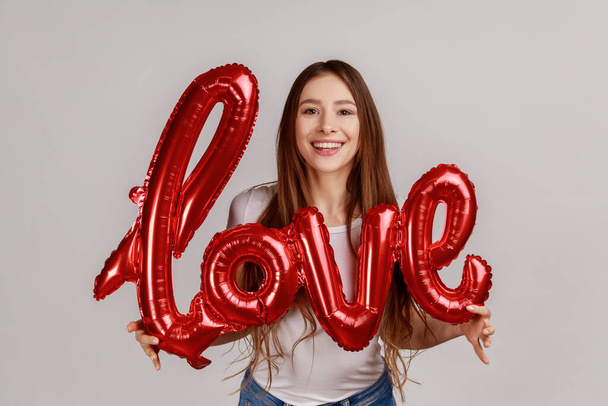 Portrait of beautiful satisfied romantic woman holding foil balloon letters, looking at camera, expressing love, wearing white T-shirt. Indoor studio shot isolated on gray background. - Photo, image