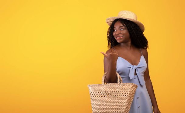 ストローバッグと帽子を持つアフリカ系アメリカ人女性は、オレンジのスタジオの背景の上に空白のスペースを指摘し、バナーデザイン - 写真・画像
