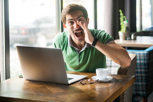 Portrait of crazy man wearing green T-shirt, sitting with hand near mouth and screaming to share something while working online on laptop, Indoor shot near big window, cafe background. - Фото, изображение