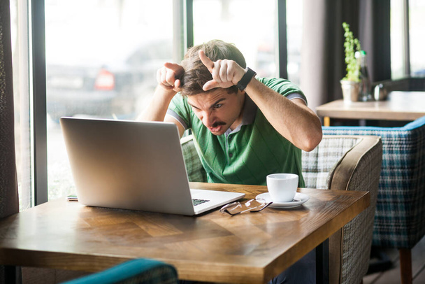 Portrait of angry businessman wearing green T-shirt at work, looking at laptop monitor, showing bull horn gesture, threatening to attack. Indoor shot near big window, cafe background. - Foto, immagini