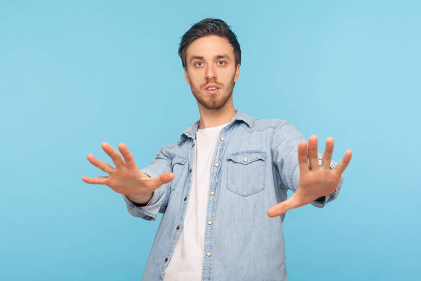 Portrait of frightened hysterical attractive man wearing denim shirt, standing, raised hands to defend from fear, horrified and shocked. Indoor studio shot isolated on blue background. - Foto, imagen