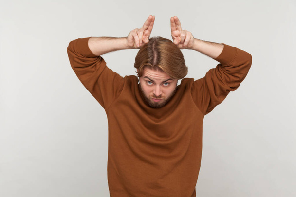 Portrait of angry bully man with beard wearing sweatshirt, showing bull horn gesture with fingers over head, looking hostile and threatening. Indoor studio shot isolated on gray background. - Zdjęcie, obraz