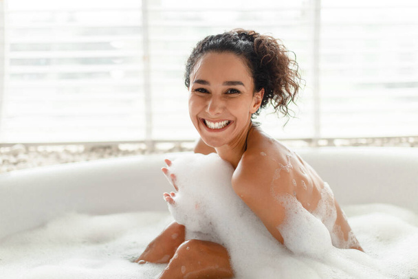 Neşeli genç kadın portresi köpükle banyo yapıyor, kameraya gülümsüyor, sabahları sıcak küvette dinleniyor. - Fotoğraf, Görsel