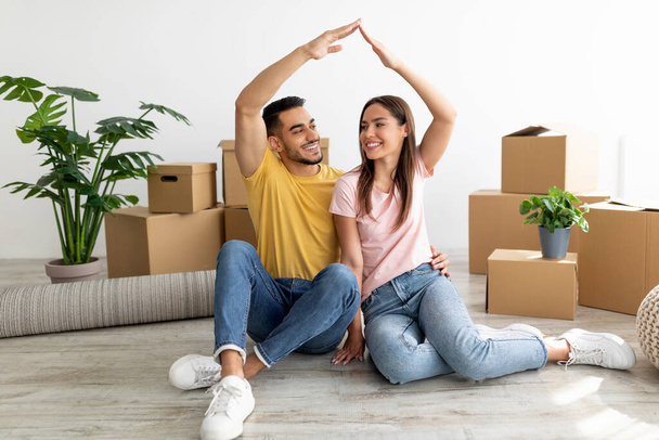 Happy millennial international couple making house gesture, sitting on floor among carton boxes, moving to new apartment - Foto, immagini