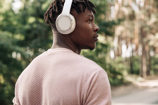 Active healthy happy African American runner, listening to music with headphones, jogging outdoors on the road in the forest, side view of young athletic man running in nature - Zdjęcie, obraz