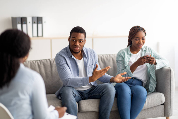 Раздражённый чёрный парень жалуется на зависимость своих жён от смартфонов во время психологической консультации в офисе - Фото, изображение