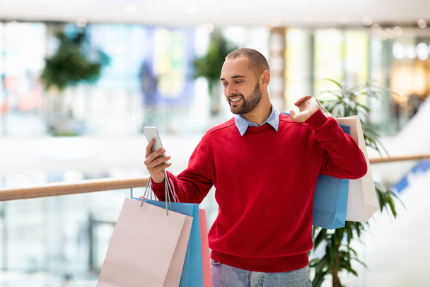 Jeune homme positif avec des sacs-cadeaux en utilisant un téléphone portable au centre commercial, en prenant un selfie ou une photo, en ayant un appel vidéo, un espace libre - Photo, image