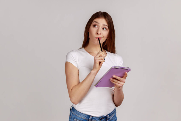 Thoughtful woman standing with paper notebook and pen, looking away with pensive expression, making to do list, wearing white T-shirt. Indoor studio shot isolated on gray background. - Foto, imagen